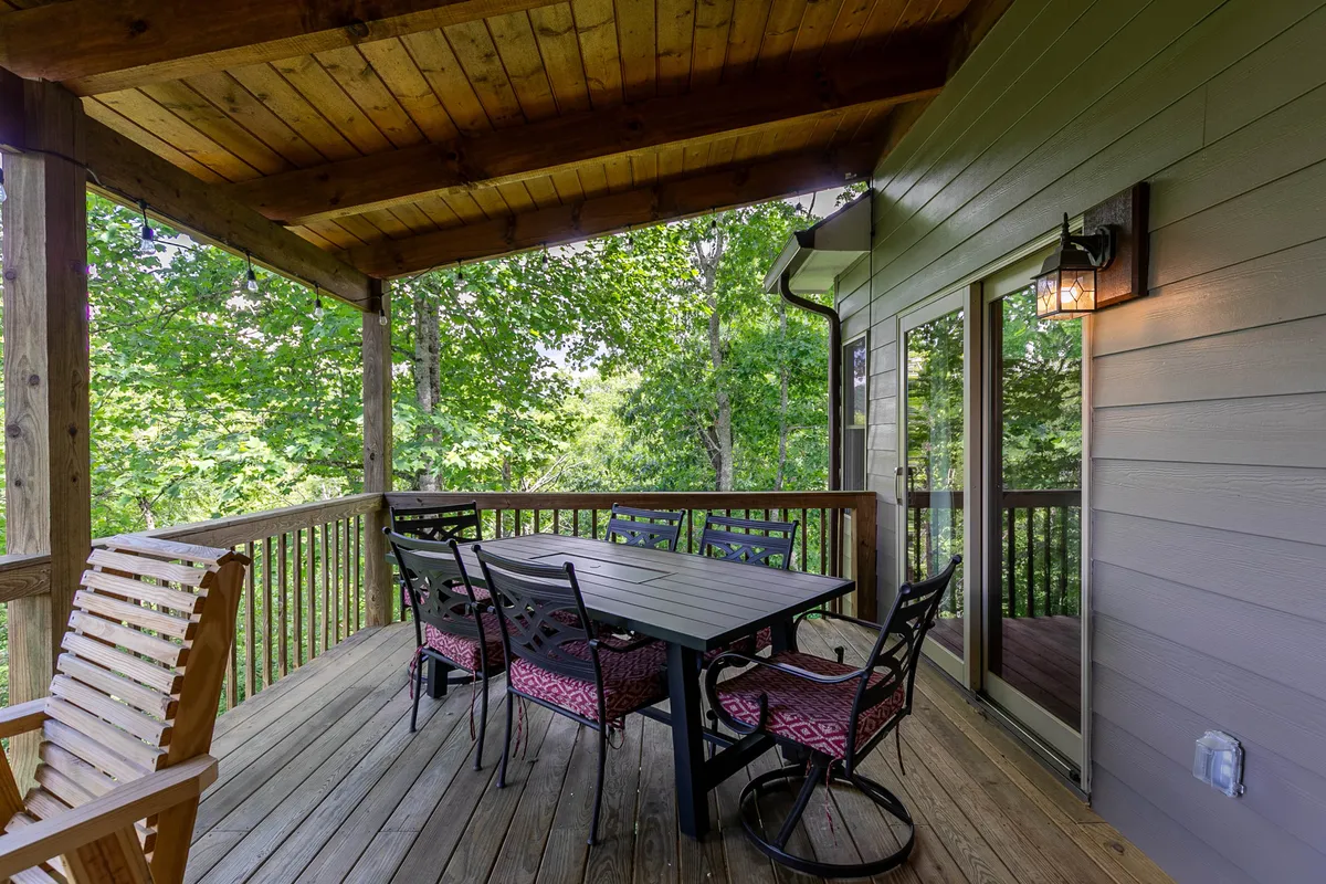A covered deck with a large outdoor dining table and a wooden rocking chair, surrounded by lush greenery. The tranquil setting offers plenty of seating for outdoor meals or relaxing with a book, with hints of mountain views beyond.