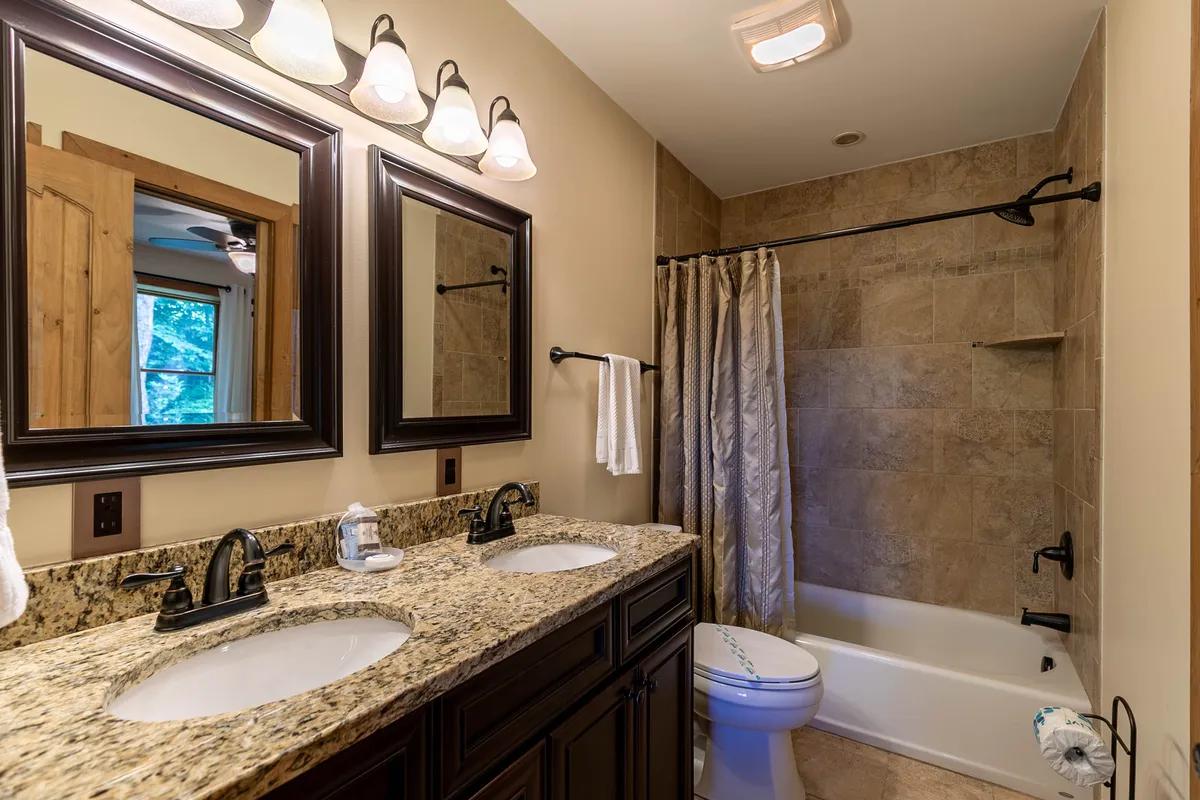 This bathroom features a double vanity with granite countertop, framed mirrors, a bathtub, and elegant light fixtures, providing a calm and functional retreat just steps from the lounge.