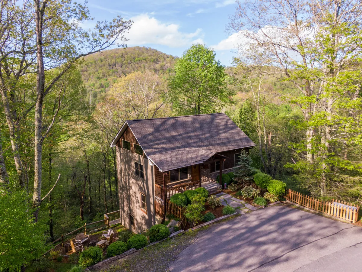 A wooden cabin nestled among lush trees with a landscaped garden and a path leading to the entrance. The scene is inviting, evoking feelings of coming home after a day of outdoor activity. The porch provides a place to relax and enjoy the peaceful, natural setting.