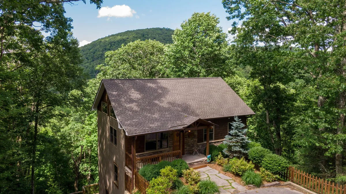 The image showcases the exterior of a charming single-family home nestled among lush trees, with a welcoming porch and a pathway leading through the serene wooded landscape. The setting evokes a sense of peace and connection to nature, highlighting the tranquility and seclusion of the property.