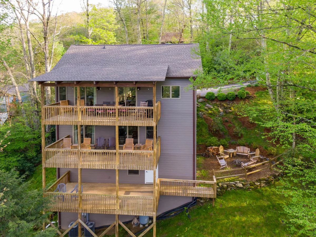 A wide exterior shot reveals the home’s expansive wooden decks on multiple levels, with large windows that draw in mountain light. In the foreground, a cozy fire pit area with seating is surrounded by lush greenery, offering a clear sense of the home’s seamless indoor-outdoor flow and its readiness for year-round gatherings.