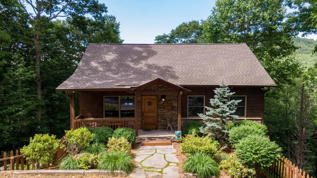 A welcoming exterior shot of the home's stone facade with a wooden front door, surrounded by lush greenery and a pathway leading up to the house. The porch invites relaxation, and the overall setting feels both inviting and central to the local area.