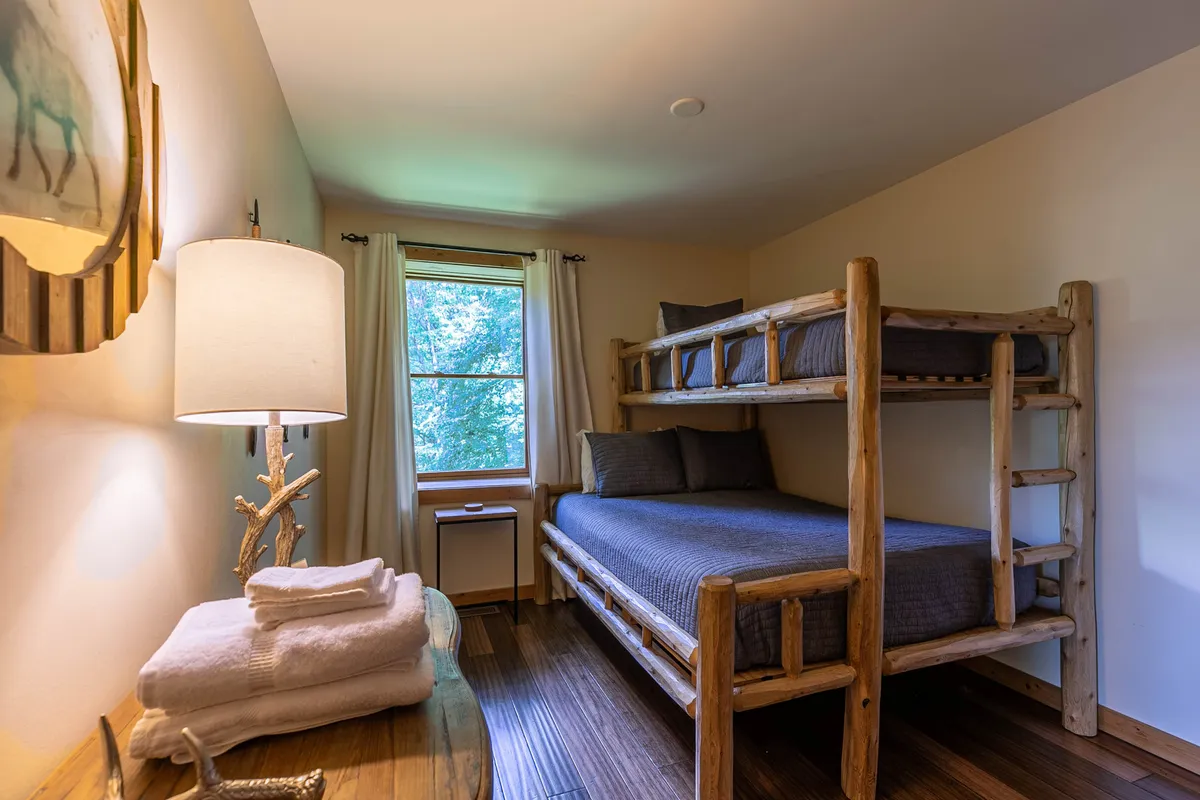 A comfortable bedroom with a twin-over-queen bunk bed, a lamp, a window with natural light, and a wooden table nearby. The layout is perfect for flexible sleeping arrangements, ideal for families or groups.
