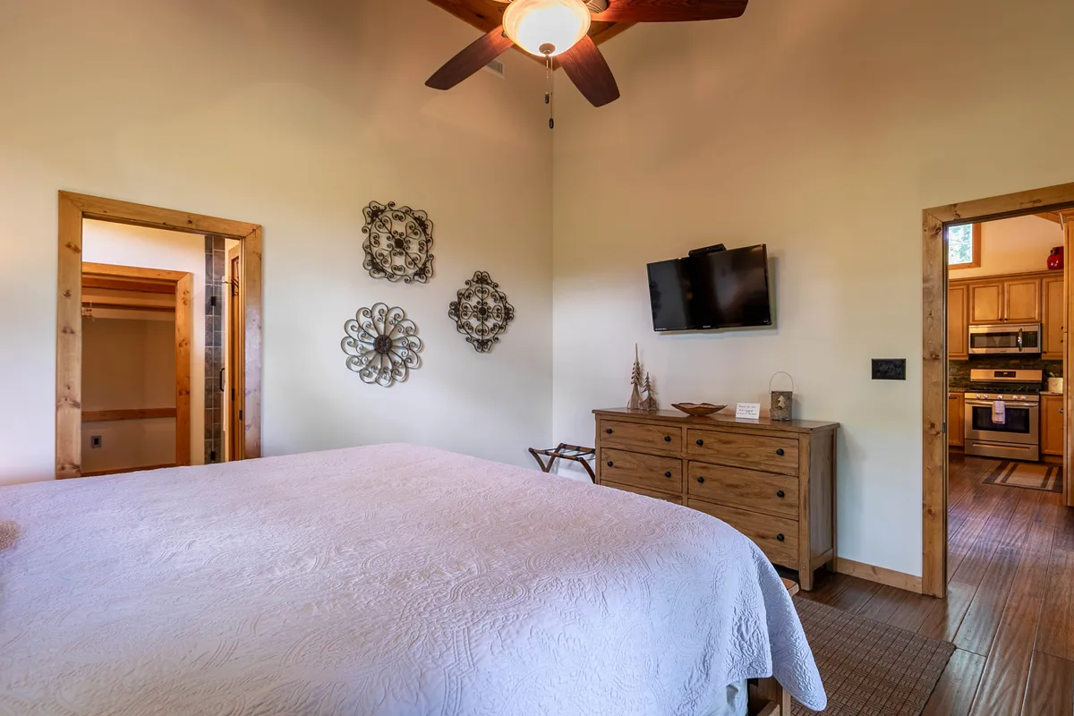 A tranquil bedroom displaying a bed with a white quilted cover, a wooden dresser, a television, and tasteful wall art. The room receives natural light and exudes a cozy, restful vibe.