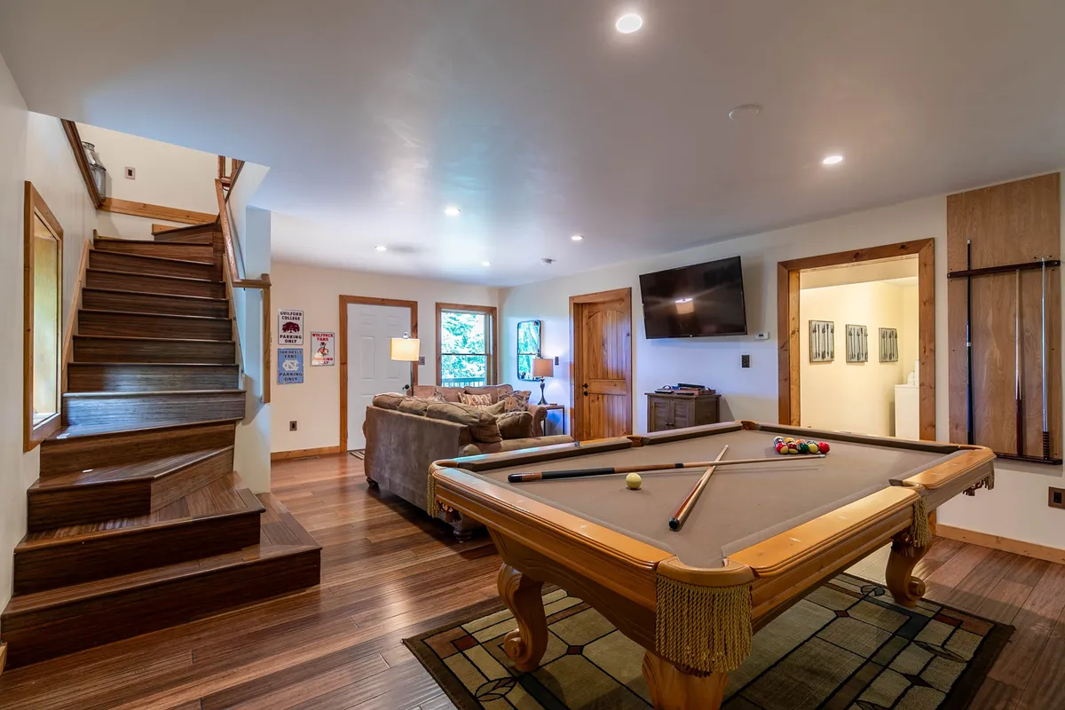 This image captures a lively billiards lounge with a pool table as the centerpiece, surrounded by a comfortable sofa, staircase, television, and decorative wall art. The space exudes a casual and friendly vibe ideal for both games and socializing.