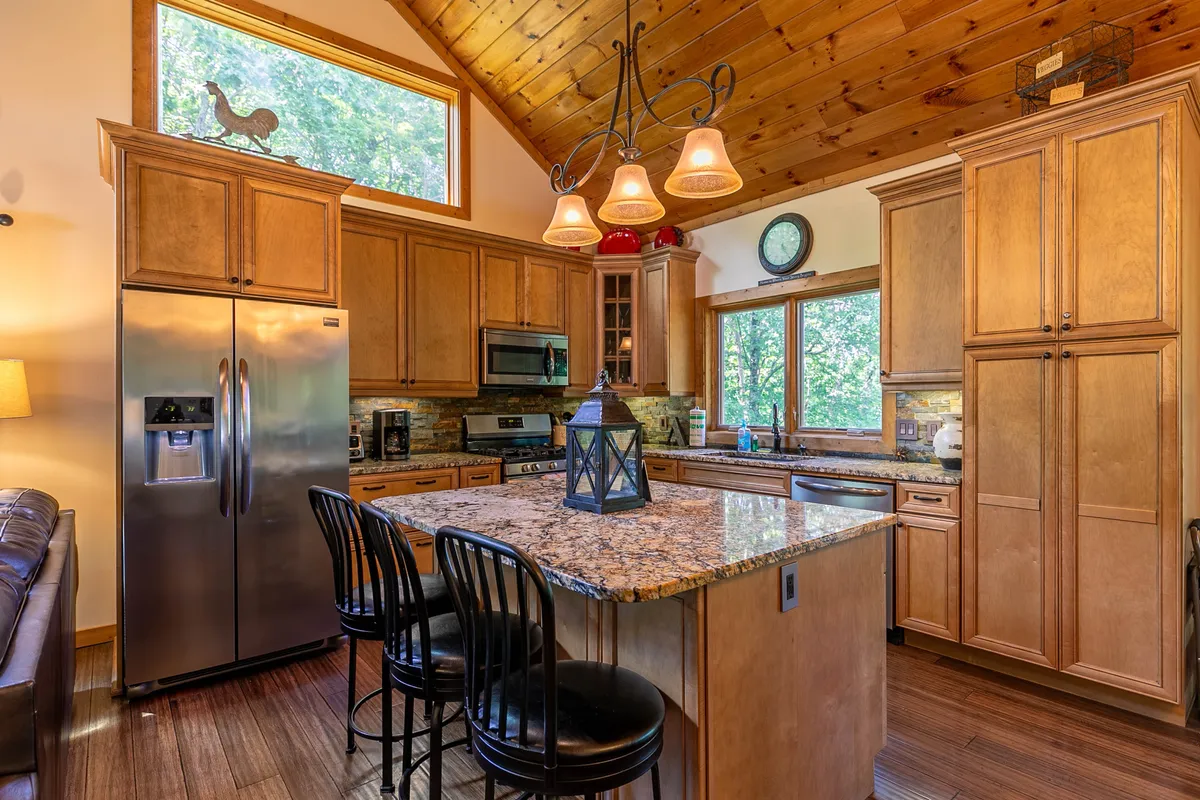 A modern gourmet kitchen with granite countertops, stainless steel refrigerator, wooden cabinetry, and pendant lighting. Large windows allow sunlight to stream in, creating an inviting space for cooking and socializing.