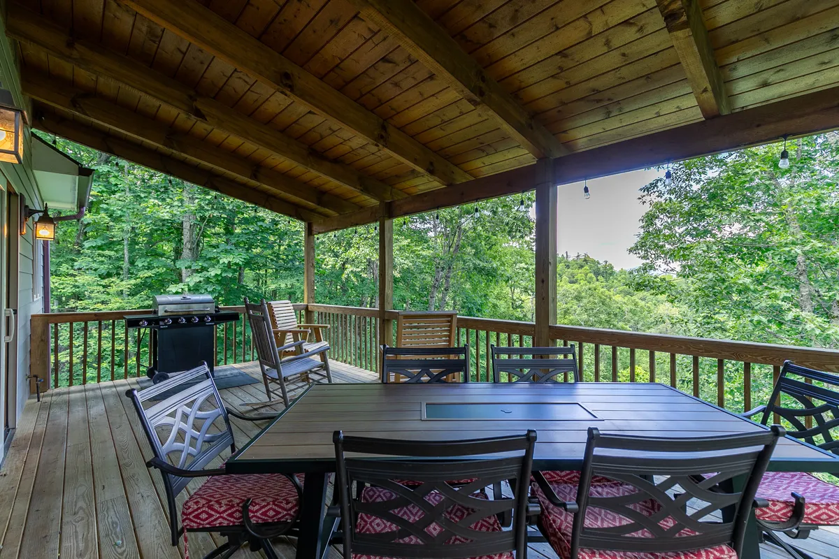A close-up of an outdoor dining table and grill nestled under wooden beams, surrounded by greenery. This inviting space is set for al fresco meals and gatherings, highlighting the home’s capacity for outdoor dining throughout the year.