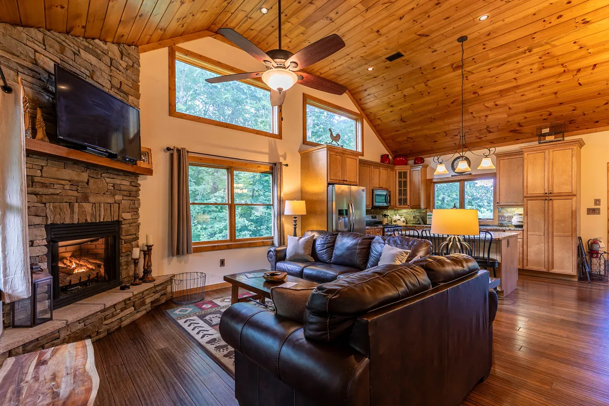 A spacious open-concept living area featuring a stunning stacked stone fireplace as the focal point. Large windows fill the space with natural light, and comfortable seating areas invite relaxation and gatherings. The kitchen area is visible in the background, seamlessly integrated with the living space.