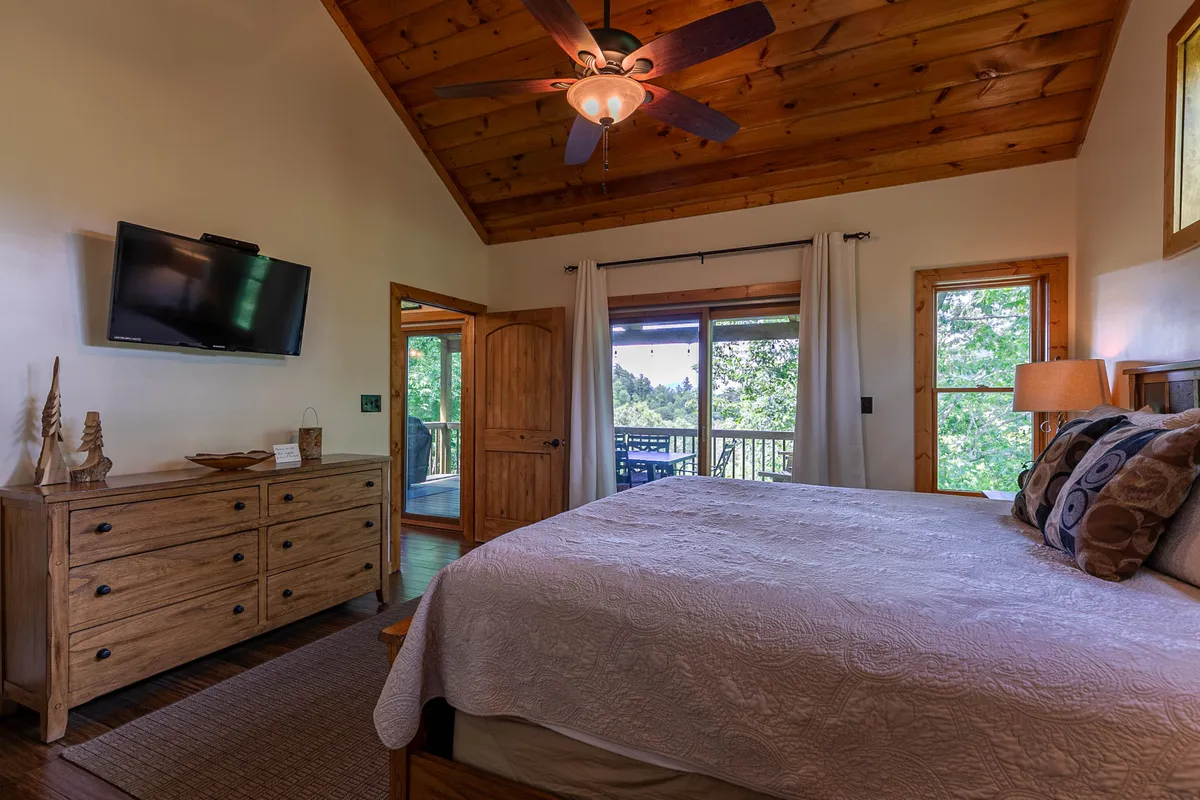 A spacious, light-filled bedroom featuring a large bed with decorative pillows, a wooden dresser, a TV mounted on the wall, and expansive windows that reveal beautiful views outside. The wooden ceiling and neutral tones create a serene atmosphere ideal for relaxation.