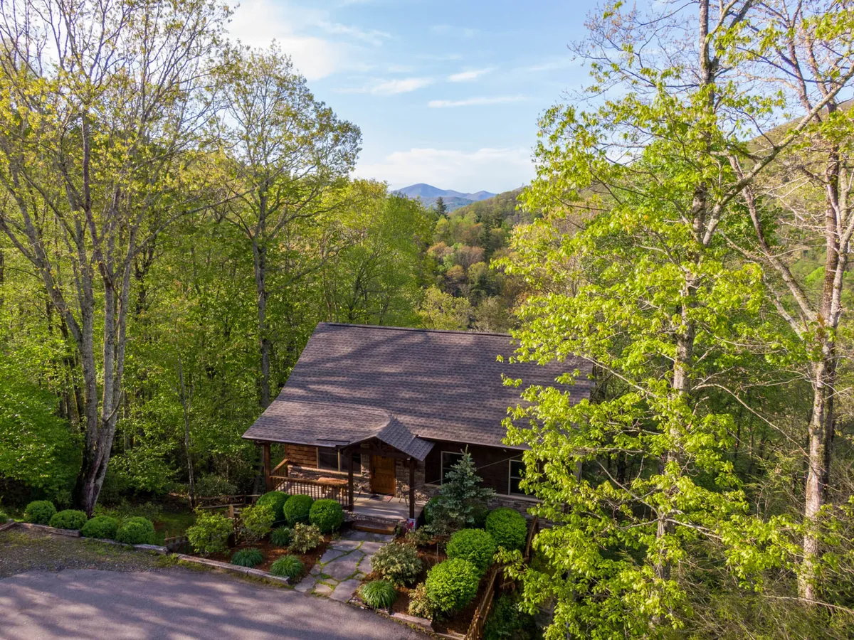 A charming log cabin with a stone pathway set amidst lush greenery and with mountains in the distance. The spacious porch suggests a welcoming spot for relaxation, nature watching, or enjoying time together outdoors.