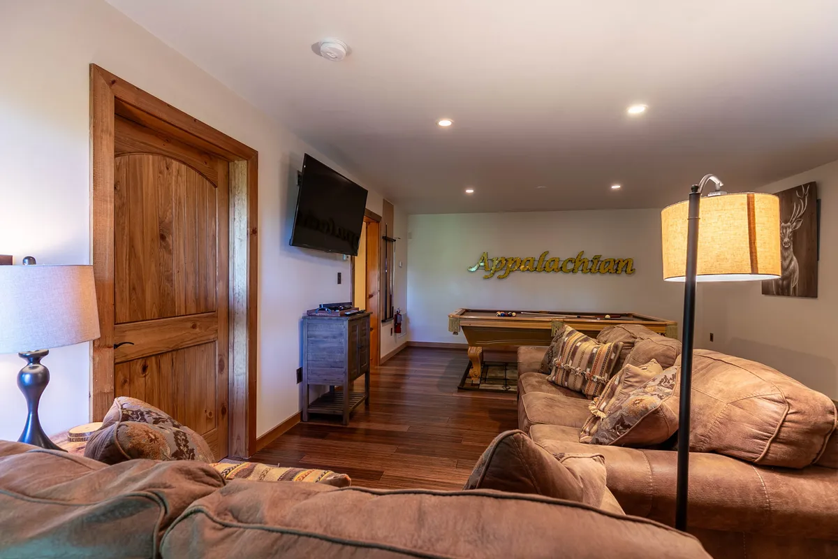 Another view of the game room highlights the pool table, a large sofa, a 'Appalachian' decorative sign, and a warm lamp, emphasizing a welcoming atmosphere perfect for both kids and adults to relax or play.